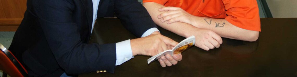 A jail chaplain wearing a dark navy blue suit is ministering to an inmate wearing an orange jumpsuit.