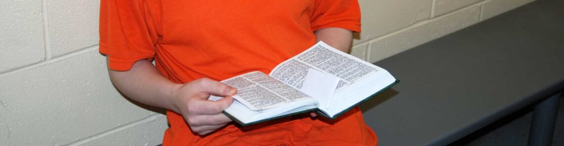 An inmate wearing an orange jumper is reading the Bible. The image is focused on the inmate's hands and their face is not visible.