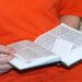 An inmate wearing an orange jumper is reading the Bible. The image is focused on the inmate's hands and their face is not visible.